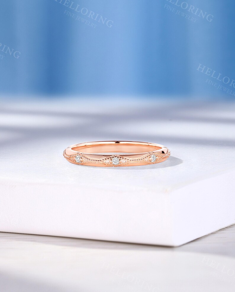 May include: A rose gold band ring featuring a series of small, round, clear stones set within a delicate, engraved metal pattern. The ring has a polished finish and a slim profile, showcasing intricate detailing along the outer surface.