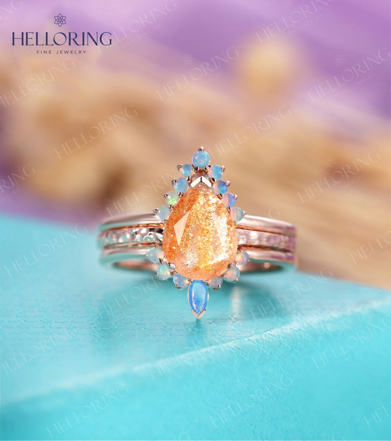May include: A pear-shaped sunstone ring with a halo of smaller opals set in a rose gold band. The ring is on a turquoise blue surface.