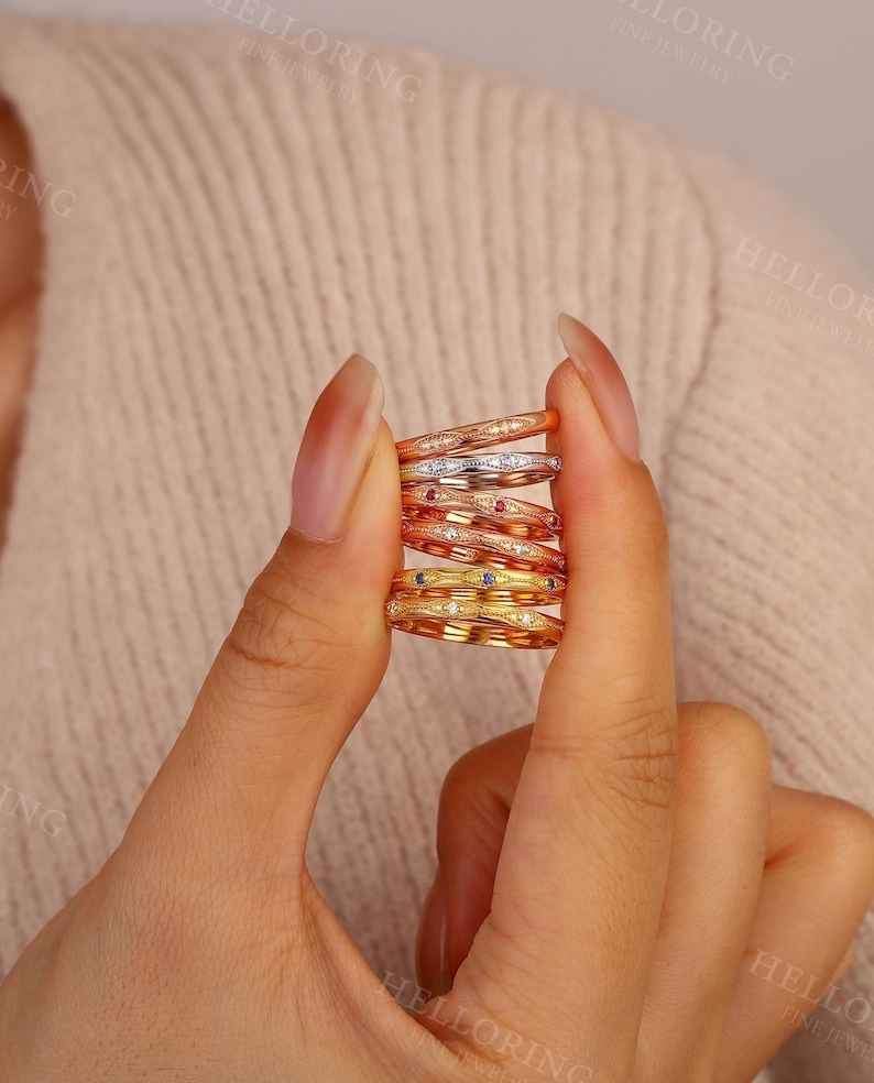 May include: A stack of six thin metal rings featuring a repeating marquise-shaped pattern with milgrain detailing. The rings are crafted in various gold tones, including yellow, rose, and white, and are accented with small embedded gemstones.
