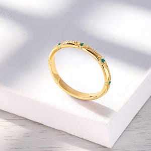 May include: A gold band ring featuring a textured, milgrain-detailed surface with small, evenly spaced, round-cut green gemstones embedded along the outer circumference. The metal has a polished finish, and the design includes repeating geometric patterns.