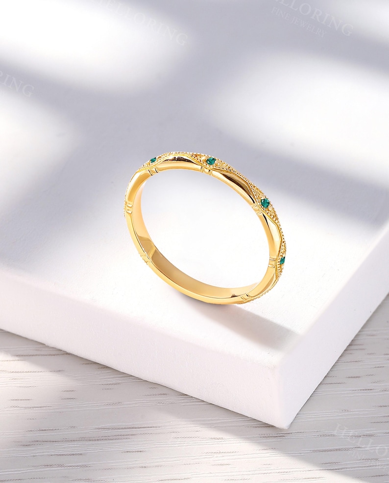 May include: A gold band ring featuring a textured, milgrain-detailed surface with small, evenly spaced, round-cut green gemstones embedded along the outer circumference. The metal has a polished finish, and the design includes repeating geometric patterns.