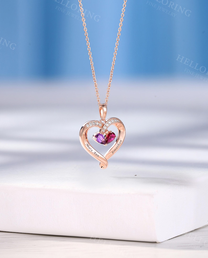 May include: Rose gold heart pendant necklace with a delicate chain. The pendant features a heart-shaped design with a smaller heart inside, holding two gemstones. The names "Mary Ann" and "Jonathan" are engraved on the pendant.