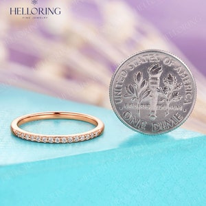 May include: Rose gold band ring with small, clear gemstones set along the top. A US dime is next to the ring for size comparison. The ring is on a light blue surface, with a blurred purple and white background. The ring is from Helloring Fine Jewelry.