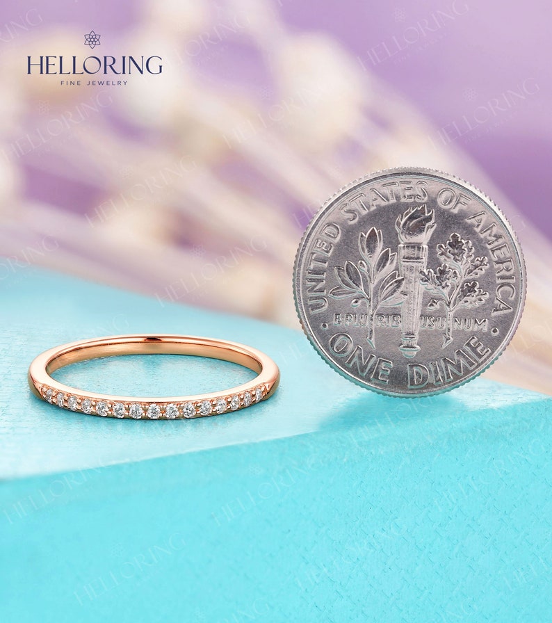 May include: Rose gold band ring with small, clear gemstones set along the top. A US dime is next to the ring for size comparison. The ring is on a light blue surface, with a blurred purple and white background. The ring is from Helloring Fine Jewelry.
