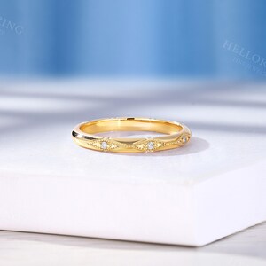 May include: A yellow gold band ring featuring a series of repeating marquise-shaped engravings along the outer surface. Each engraved section contains a small, round, clear gemstone set in the center, accented by delicate milgrain detailing.