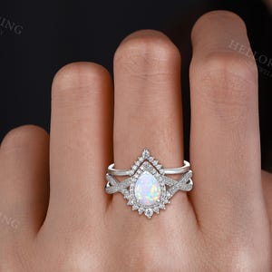 May include: A two-piece silver ring set. The engagement ring features a pear-shaped opal center stone surrounded by a halo of small diamonds. The band has a twisted design. The wedding band is a simple silver band with small diamonds.