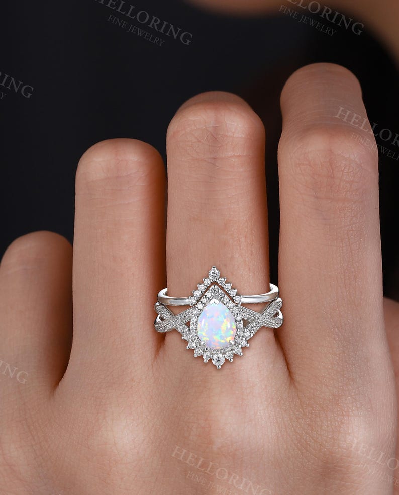 May include: A two-piece silver ring set. The engagement ring features a pear-shaped opal center stone surrounded by a halo of small diamonds. The band has a twisted design. The wedding band is a simple silver band with small diamonds.