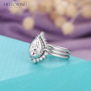May include: A pear-shaped diamond engagement ring with a halo setting and a delicate band. The ring is set on a light blue background.