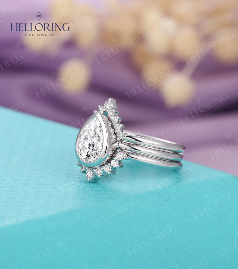 May include: A pear-shaped diamond engagement ring with a halo setting and a delicate band. The ring is set on a light blue background.