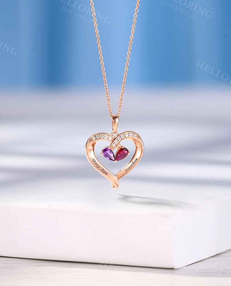May include: Rose gold heart pendant necklace with a fine chain. The pendant has two gemstones, one purple and one red, and is decorated with small clear stones. The names "Mary Ann" and "Jonathan" are engraved on the heart.