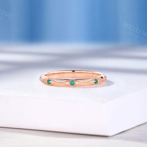 May include: A rose gold band ring featuring a series of small, round, vibrant green gemstones set at intervals. The metal surface displays intricate milgrain detailing and a repeating engraved pattern along the entire circumference of the band.