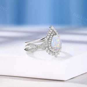 May include: A silver engagement ring with a pear-shaped opal gemstone. The opal is surrounded by a halo of small, sparkling diamonds. The ring band features a delicate, intertwined design.