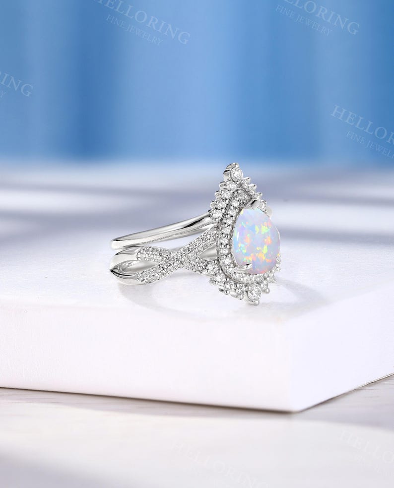 May include: A silver engagement ring with a pear-shaped opal gemstone. The opal is surrounded by a halo of small, sparkling diamonds. The ring band features a delicate, intertwined design.