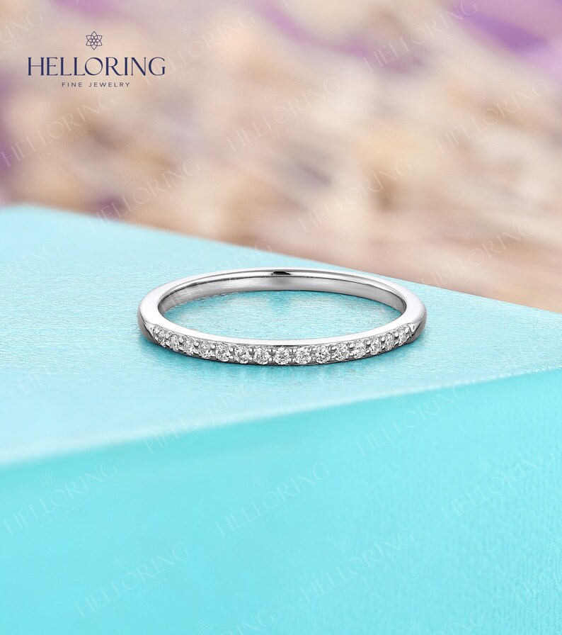 May include: A delicate silver ring with a band of small, clear gemstones. The ring is set against a light blue surface, with a blurred background of purple and white. The ring is a fine jewelry item.