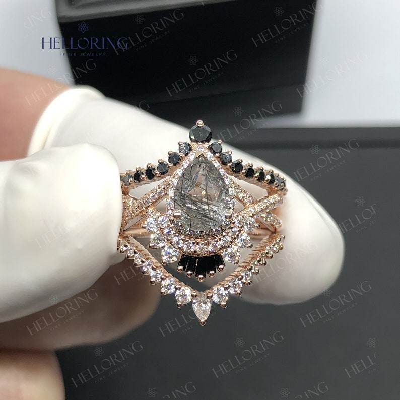 May include: A pear-shaped engagement ring with a grey and black gemstone surrounded by a halo of smaller diamonds. The ring is set in rose gold and features a delicate band with black diamonds.