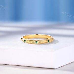 May include: A gold band ring featuring a series of repeating marquise-shaped engravings, each containing a small, round, vibrant green gemstone. The metal surface displays a polished finish with intricate milgrain detailing along the edges.