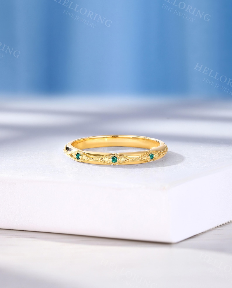 May include: A gold band ring featuring a series of repeating marquise-shaped engravings, each containing a small, round, vibrant green gemstone. The metal surface displays a polished finish with intricate milgrain detailing along the edges.