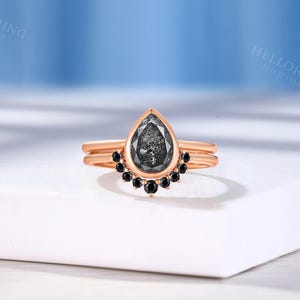 以下が含まれることがあります： 梨型のブラックダイヤモンドと、小さなブラックダイヤモンドのハローがセットされた、ローズゴールドの婚約指輪です。