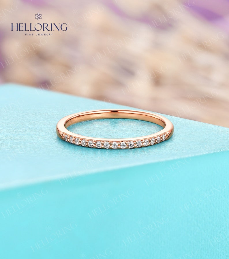 May include: Rose gold band ring with a row of small, clear gemstones. The ring is set against a light blue background. The ring is a delicate piece of jewelry.