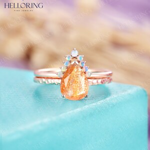 May include: A pear-shaped sunstone engagement ring set in rose gold with a halo of white opals. The ring is on a blue surface.