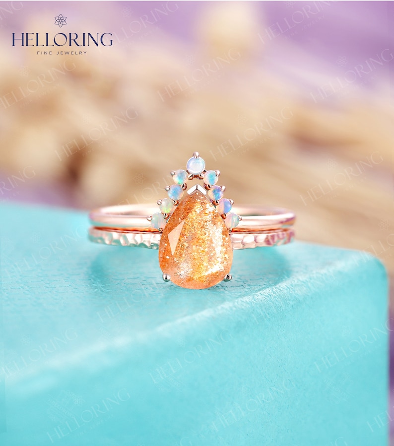 May include: A pear-shaped sunstone engagement ring set in rose gold with a halo of white opals. The ring is on a blue surface.