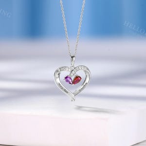 May include: A silver heart pendant necklace with a chain. The pendant has a heart design with clear stones and two gemstones, one purple and one red. The names "Mary Ann" and "Jonathan" are engraved on the pendant.