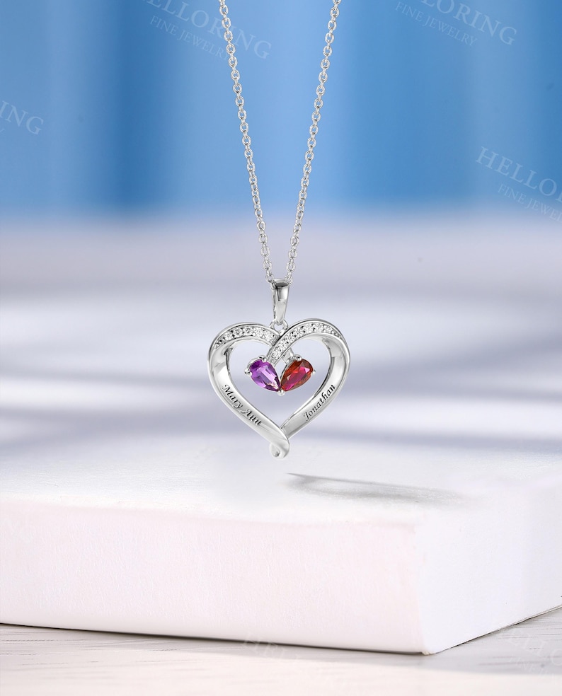 May include: A silver heart pendant necklace with a chain. The pendant has a heart design with clear stones and two gemstones, one purple and one red. The names "Mary Ann" and "Jonathan" are engraved on the pendant.