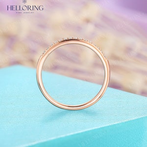 May include: Rose gold ring with small, clear gemstones set along the top. The ring is displayed against a light blue surface with a blurred background. The ring is from HELLORING FINE JEWELRY.
