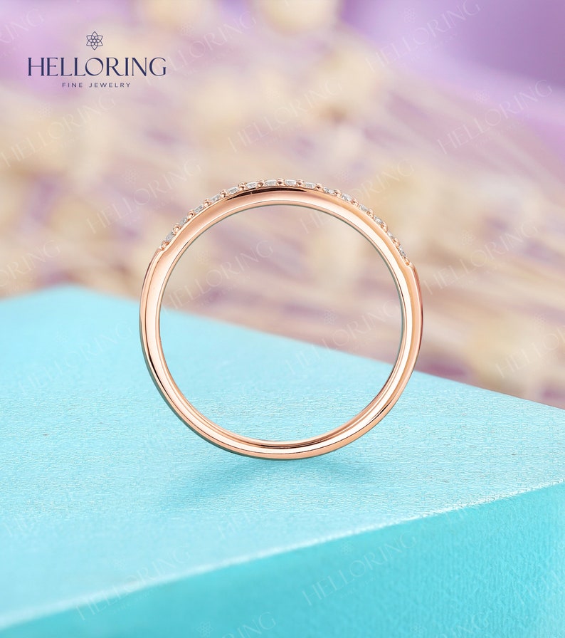 May include: Rose gold ring with small, clear gemstones set along the top. The ring is displayed against a light blue surface with a blurred background. The ring is from HELLORING FINE JEWELRY.