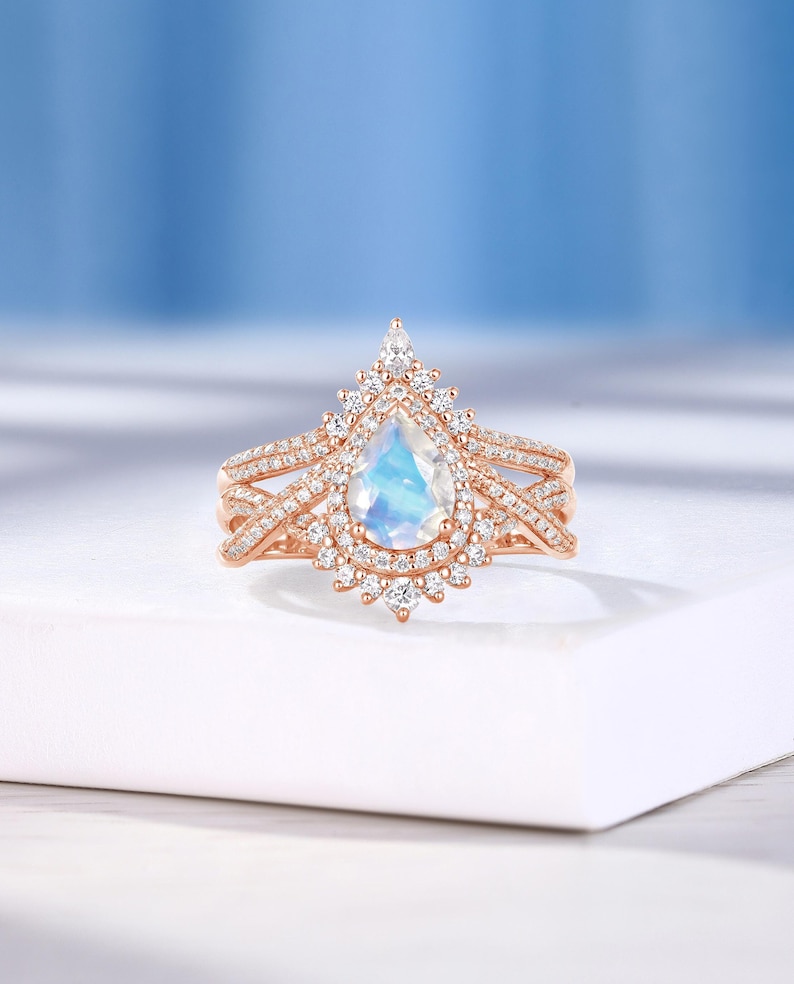 Peut inclure: Une bague de fian&ccedil;ailles en or rose avec une pierre de lune en forme de poire et un halo de petits diamants. La bague pr&eacute;sente un design d&eacute;licat et complexe avec une double bande.