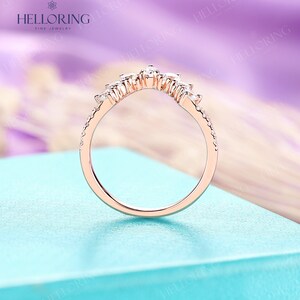 May include: A delicate rose gold wedding band with a curved design featuring a row of small diamonds. The band is on a light blue background.