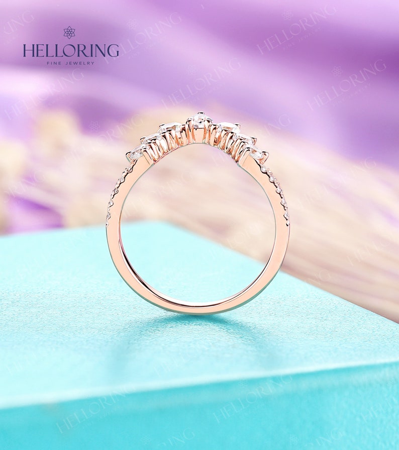 May include: A delicate rose gold wedding band with a curved design featuring a row of small diamonds. The band is on a light blue background.