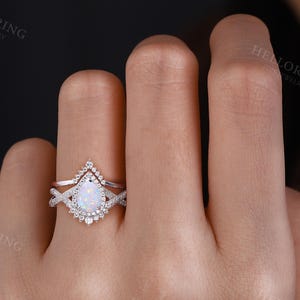May include: A pear-shaped opal engagement ring with a delicate, twisted band and a halo of smaller diamonds. The ring is set in white gold and is worn on a hand.