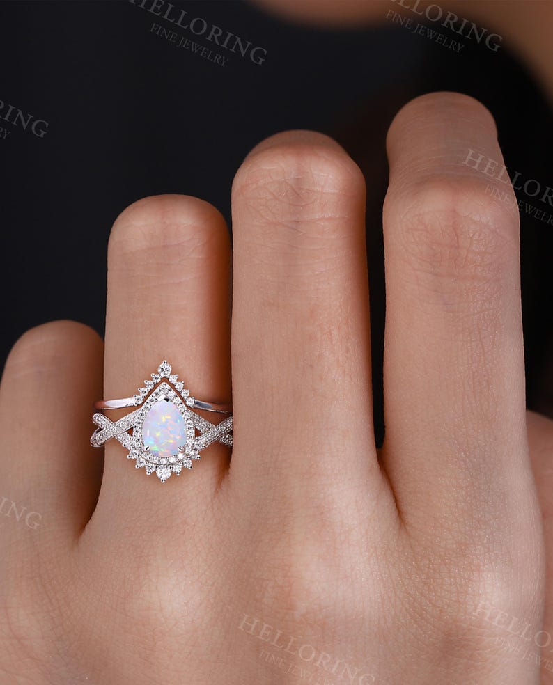 May include: A pear-shaped opal engagement ring with a delicate, twisted band and a halo of smaller diamonds. The ring is set in white gold and is worn on a hand.