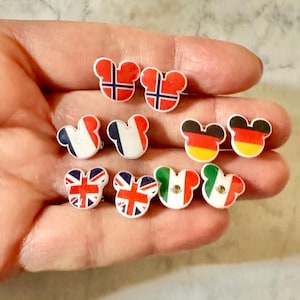 May include: A collection of small, colorful stud earrings in the shape of a mouse head. Each earring features a different flag design, including Norway, France, Germany, the United Kingdom, and Italy. The earrings are held in a hand.