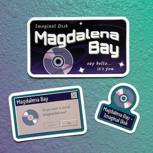 May include: Three stickers featuring a fictional computer game called "Magdalena Bay". The first sticker is a blue and purple design with a white disc and the text "Imaginal Disk Magdalena Bay say hello... it's you.". The second sticker is a blue and white window with the text "Magdalena Bay Do you want to install Imaginal Disk.exe? Cancel Yes". The third sticker is a blue and white design with a white disc and the text "Magdalena Bay - Imaginal Disk".