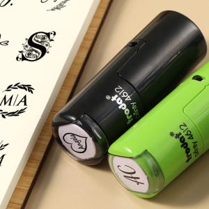 Może przedstawiać: Dwa automatyczne stemple, jeden czarny i jeden limonkowy, z napisem "trodat 4612" z boku. Poduszki stempli pokazują słowa "Angela" i "Nc" w skrypcie. Notatnik wyświetla różne projekty liter.