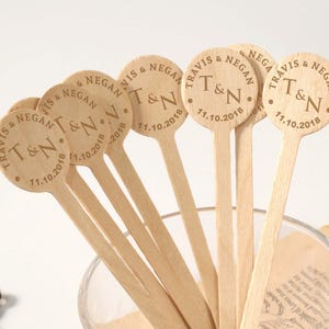 Puede incluir: Un conjunto de palitos de madera para remover bebidas con texto grabado. Cada palito tiene una parte superior circular con los nombres "Travis & Negan", las iniciales "T&N" y la fecha "11.10.2018".