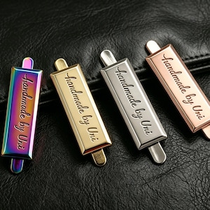 May include: Four metal tags in iridescent, gold, silver, and rose gold colors, each with the text "handmade by Uni" in a cursive font. The tags are rectangular with a small tab at each end, set against a black leather background.