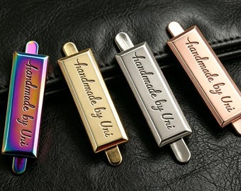 Puede incluir: Cuatro etiquetas metálicas en colores iridiscente, dorado, plateado y oro rosa, cada una con el texto "handmade by Uni" en cursiva. Las etiquetas son rectangulares con una pequeña pestaña en cada extremo, sobre un fondo de cuero negro.
