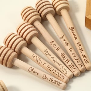 Può includere: Una collezione di cucchiaini da miele in legno chiaro, ciascuno con una testa scanalata e un manico sottile. Diversi cucchiaini sono incisi con nomi e date, suggerendo un matrimonio o un evento speciale. I cucchiaini sono disposti su una superficie bianca.