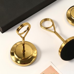 May include: Gold-colored metal place card holders with a circular base and a loop at the top. The base has a black, circular pad. The holders are arranged on a white surface, with a black box in the background.