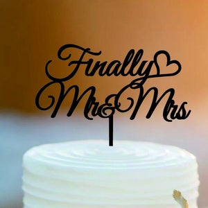 May include: A two-tiered white wedding cake with a black acrylic cake topper that reads "Finally Mr & Mrs" with a heart symbol. The cake is decorated with white frosting and a single pink rose.