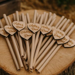 Puede incluir: Un conjunto de pajitas de papel marrón claro con adornos de madera en forma de corazón. Cada corazón está grabado con los nombres "Ashley & David". Las pajitas están dispuestas sobre una rodaja de madera, ideal para bodas.