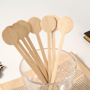 Puede incluir: Un conjunto de cucharas de madera de color claro con cabezas redondas, dispuestas en un vaso transparente. Las cucharas tienen mangos largos y delgados. El vaso est&aacute; sobre un trozo de papel con texto. Una pieza decorativa de madera est&aacute; en el fondo.