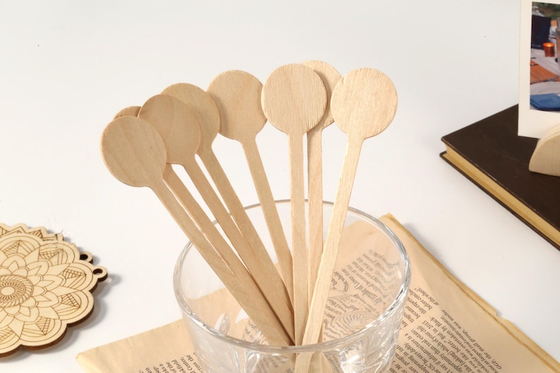 Puede incluir: Un conjunto de cucharas de madera de color claro con cabezas redondas, dispuestas en un vaso transparente. Las cucharas tienen mangos largos y delgados. El vaso est&aacute; sobre un trozo de papel con texto. Una pieza decorativa de madera est&aacute; en el fondo.