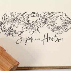 May include: A cream-colored card with a black floral design and the names "Sujadi and Hartini" in script. A wooden object with an orange floral design is in the lower left corner.