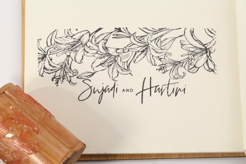 May include: A cream-colored card with a black floral design and the names "Sujadi and Hartini" in script. A wooden object with an orange floral design is in the lower left corner.