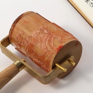 May include: A wooden patterned roller with a brass frame and handle. The roller has a floral design and is partially covered in red ink. A notepad with a floral design is in the background.