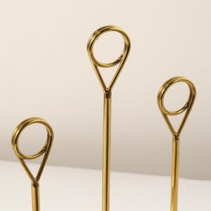 May include: Three gold-colored metal place card holders. Each holder has a slender stem that leads to a triangular shape, topped with a circular ring. The holders are arranged in a row, with varying heights.